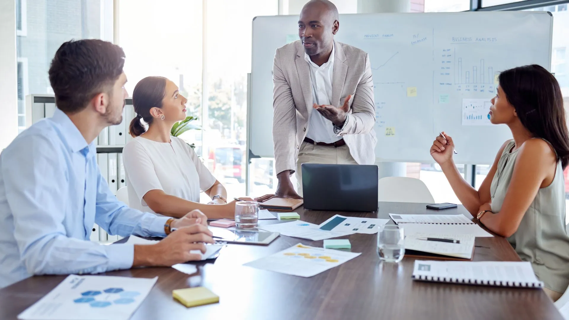 A man is standing at the head of a conference table teaching 3 other employees who are sitting around the table.