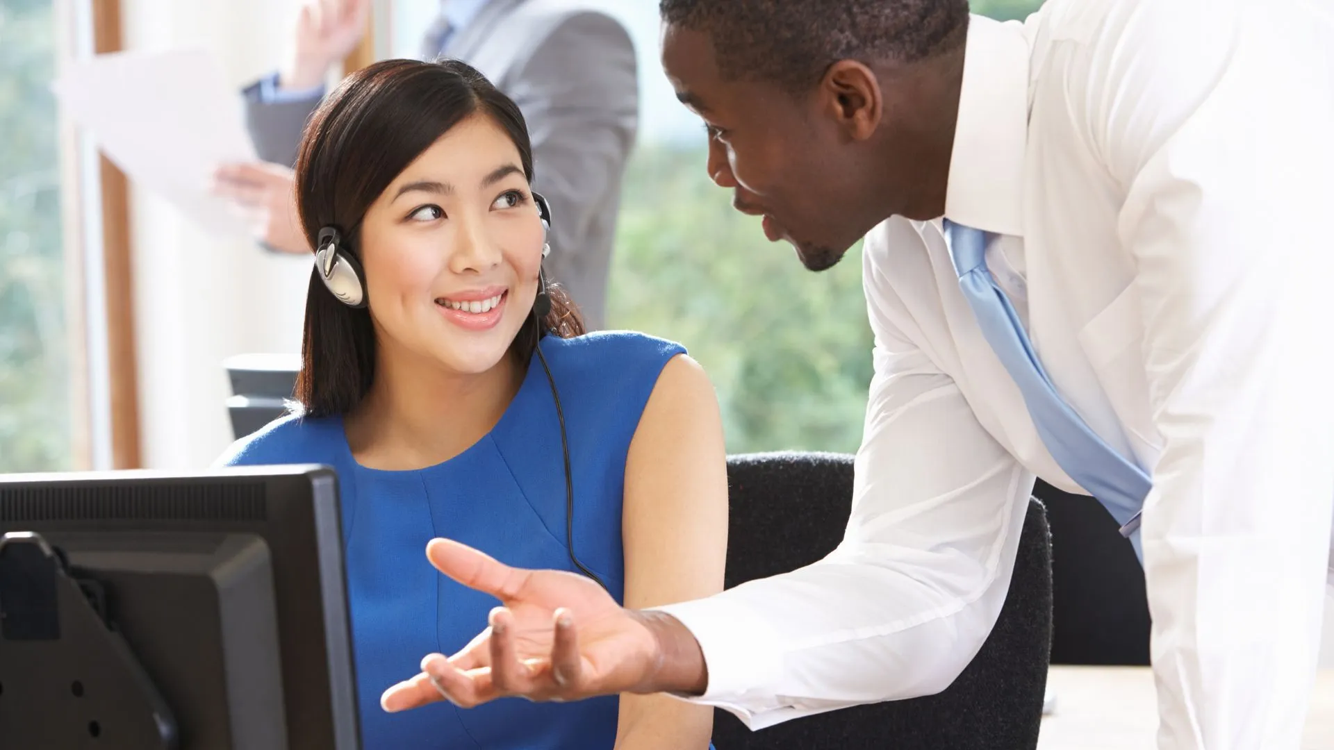 A manager is leaning over a seated employee having a conversation with them, showing them how to do something.