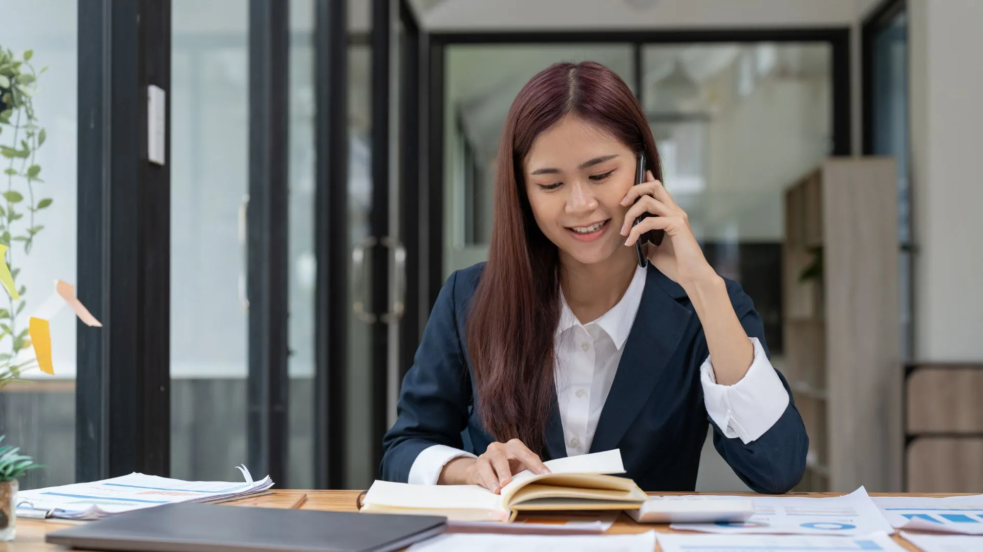 A young woman is talking on her cell phone while looking down at her notebook.