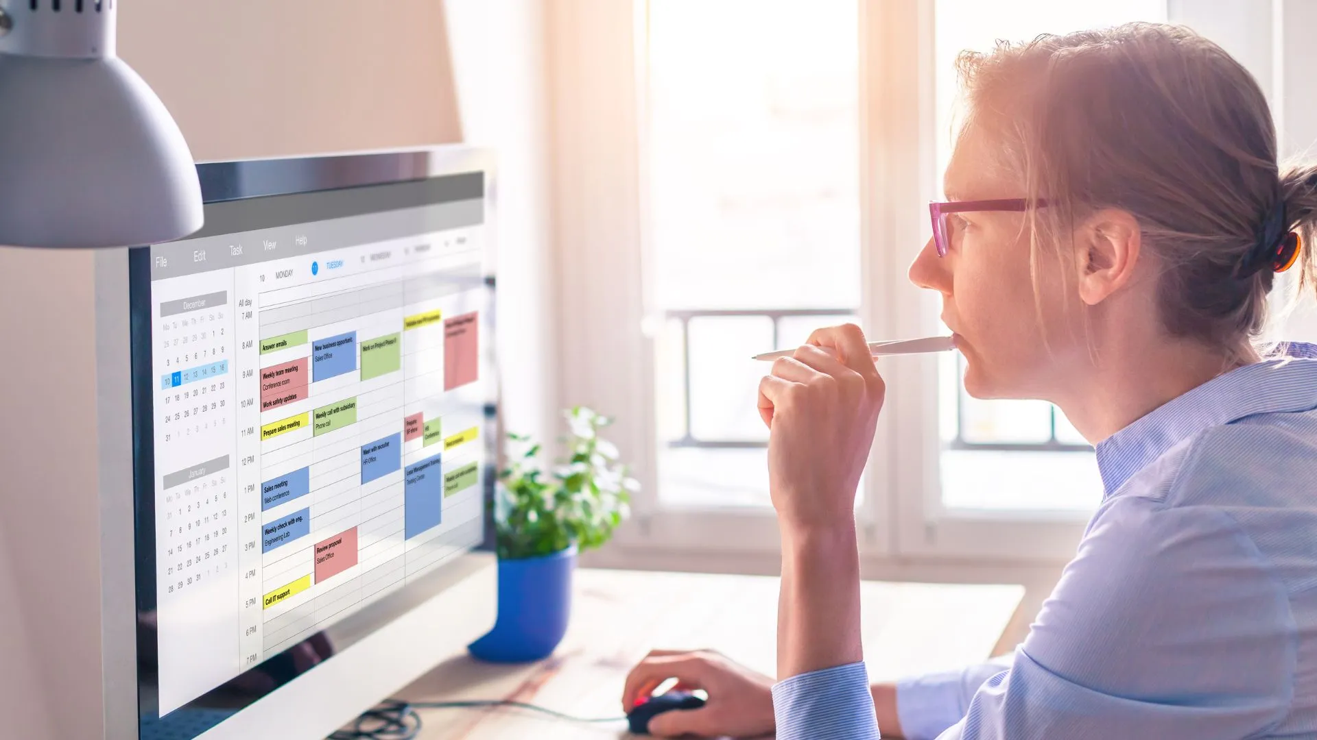 A woman in glasses is biting the end of her pen while looking at her calendar on her computer monitor. 