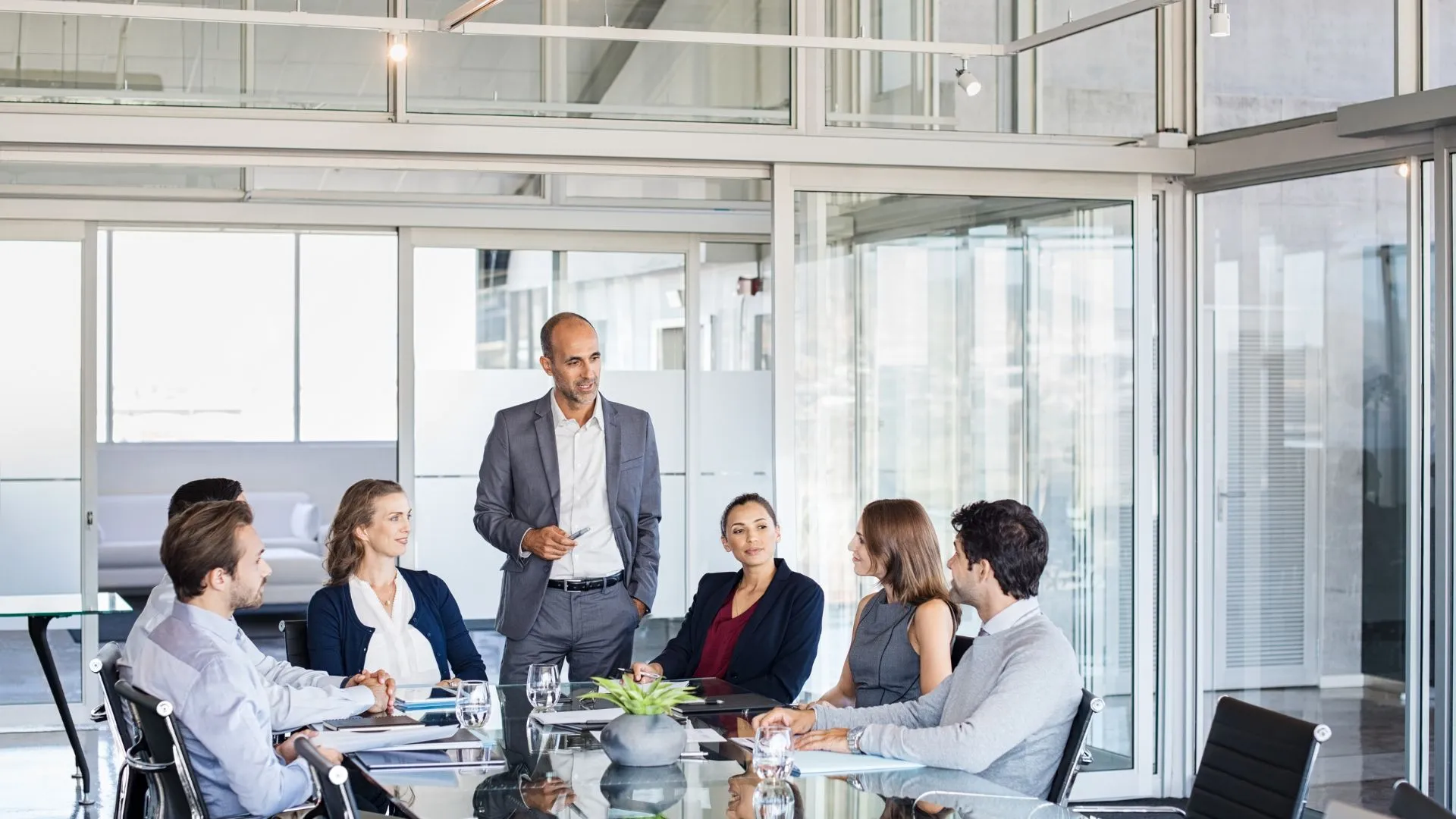 A man is standing at the head of a conference table, presenting to a diverse group of colleagues.