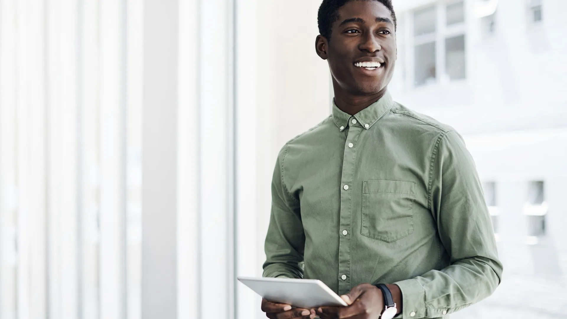 A young man is standing and holding a tablet, smiling off into the distance.