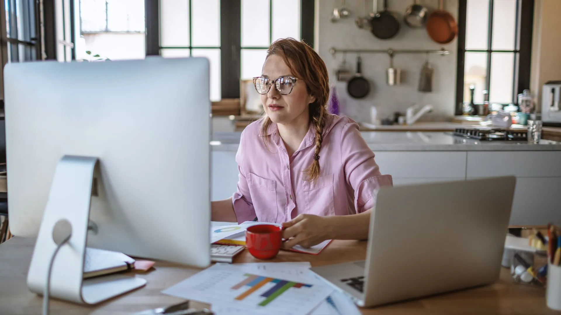 A woman is working on two computers from her kitchen table. She is holding a cup of coffee.