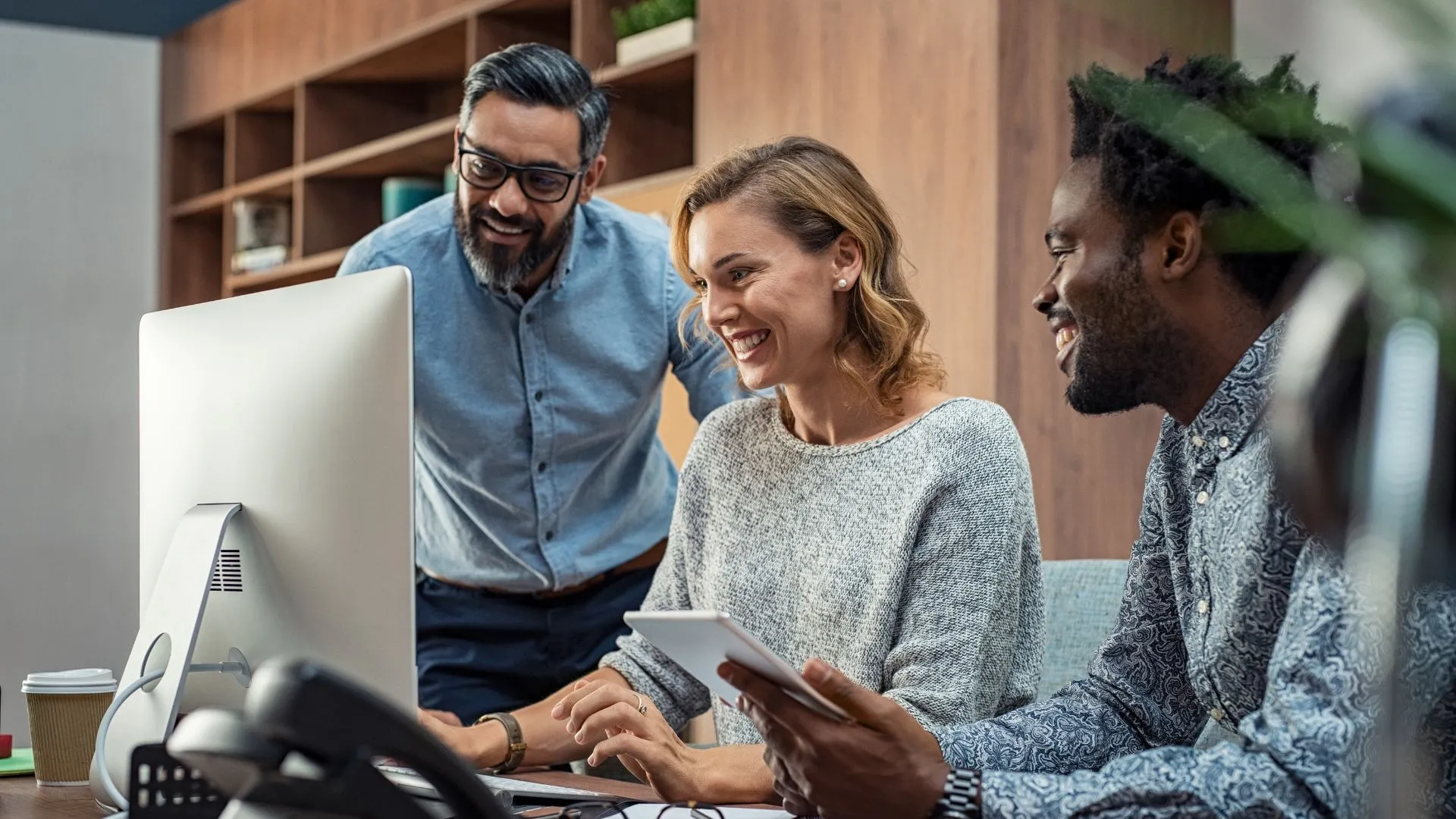 There are three people gathered around a computer looking at it and smiling. There are two men and one woman.