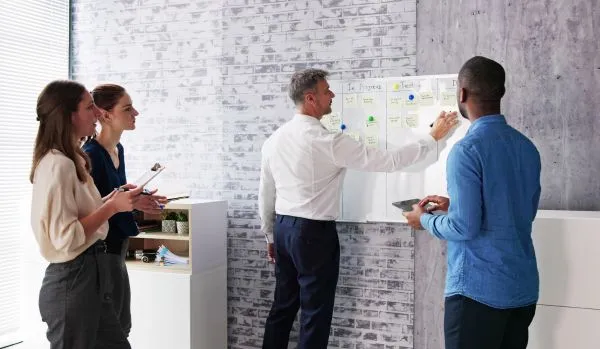 A diverse team of four people are looking at and working on a whiteboard, doing agile planning.