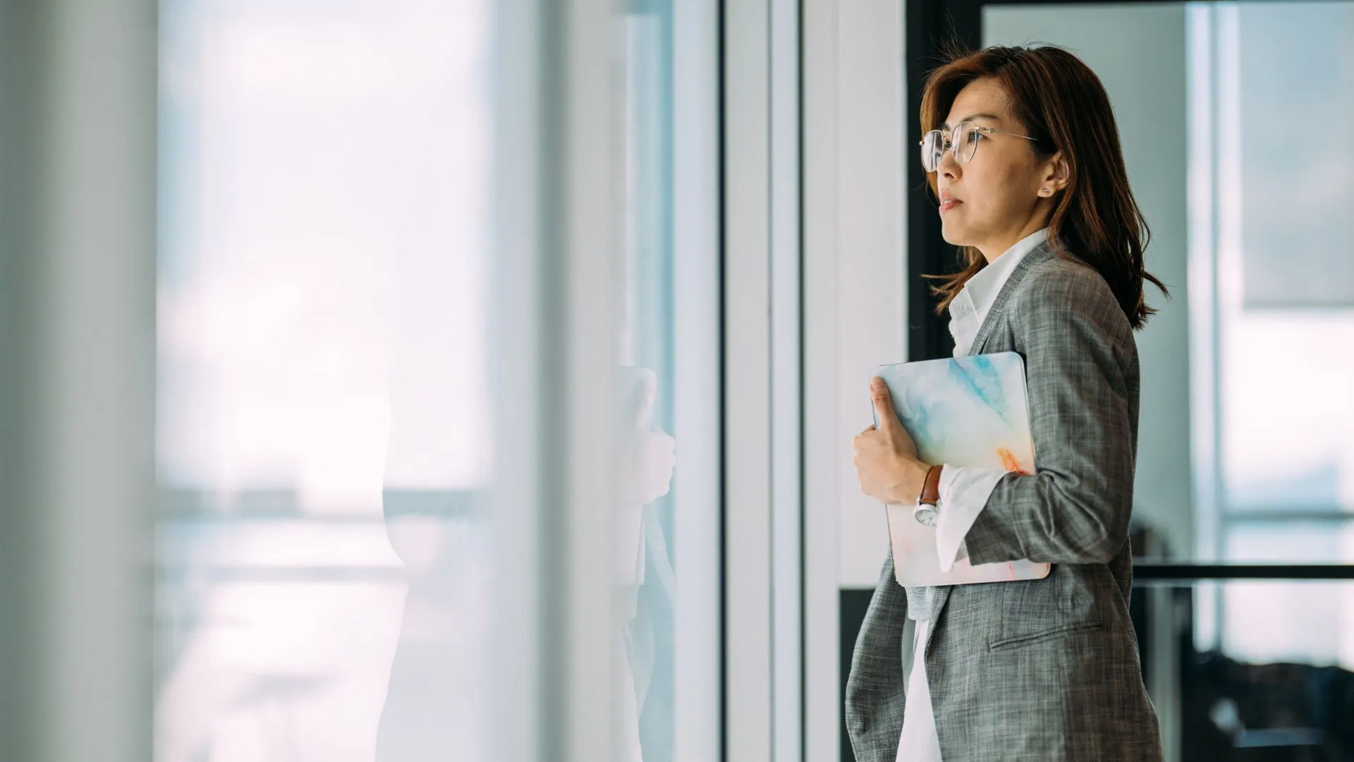 A woman wearing glasses and holding a tablet is standing and staring out the window in her office.