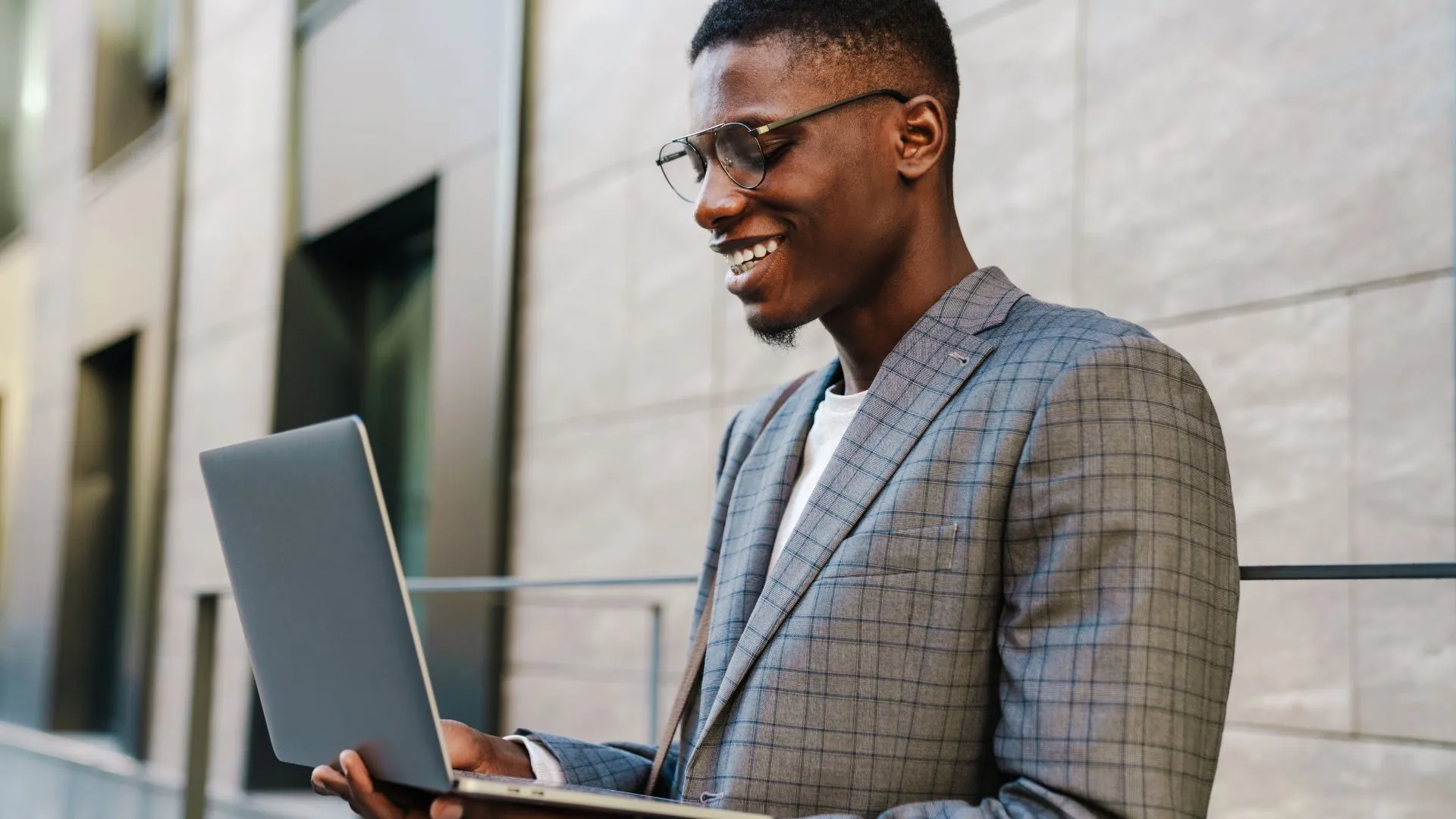A young man wearing classes is standing outside holding his laptop open and smiling down at his screen.