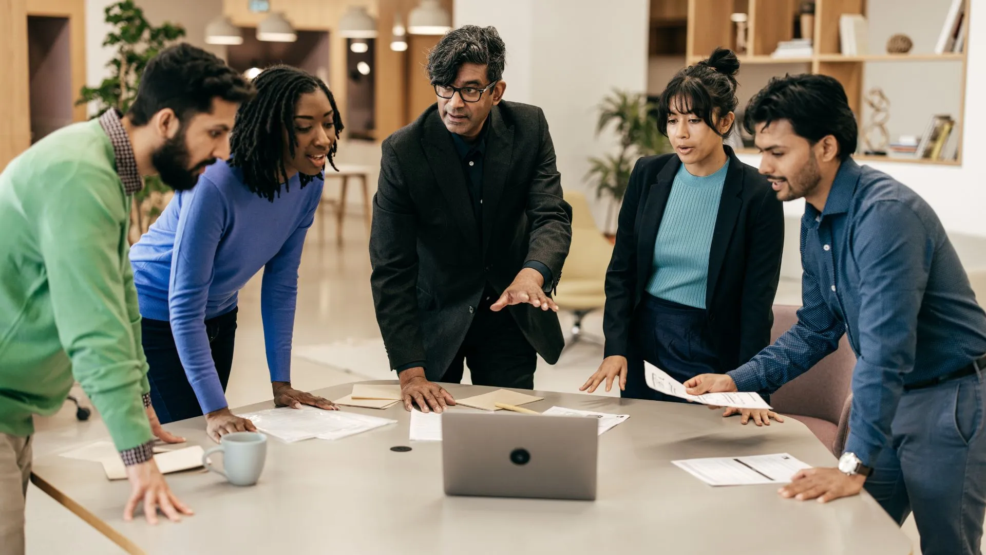 A leader is standing with his team around a table looking at a laptop, teaching them how to use AI.
