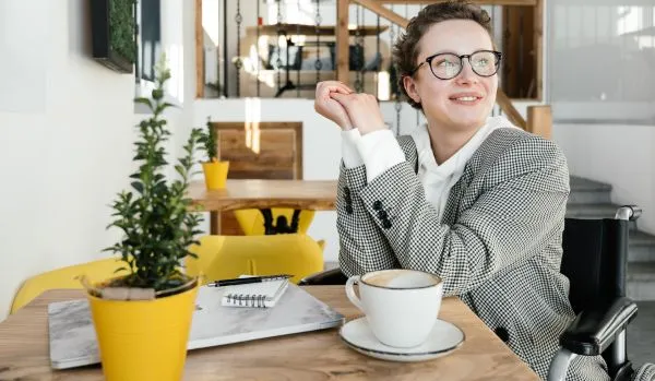 A woman in a wheelchair is sitting at a small table with her laptop and coffee, smiling into the distance.