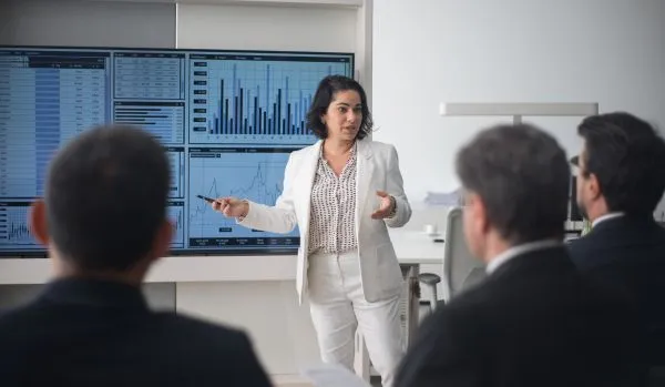 A woman in a white suit is presenting to a group of corporate executives. We are looking over the executives' shoulders.