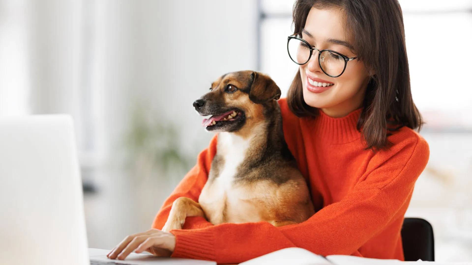 A young woman wearing glasses and an orange sweater is working on her laptop with her small dog sitting in her lap. Both are smiling.