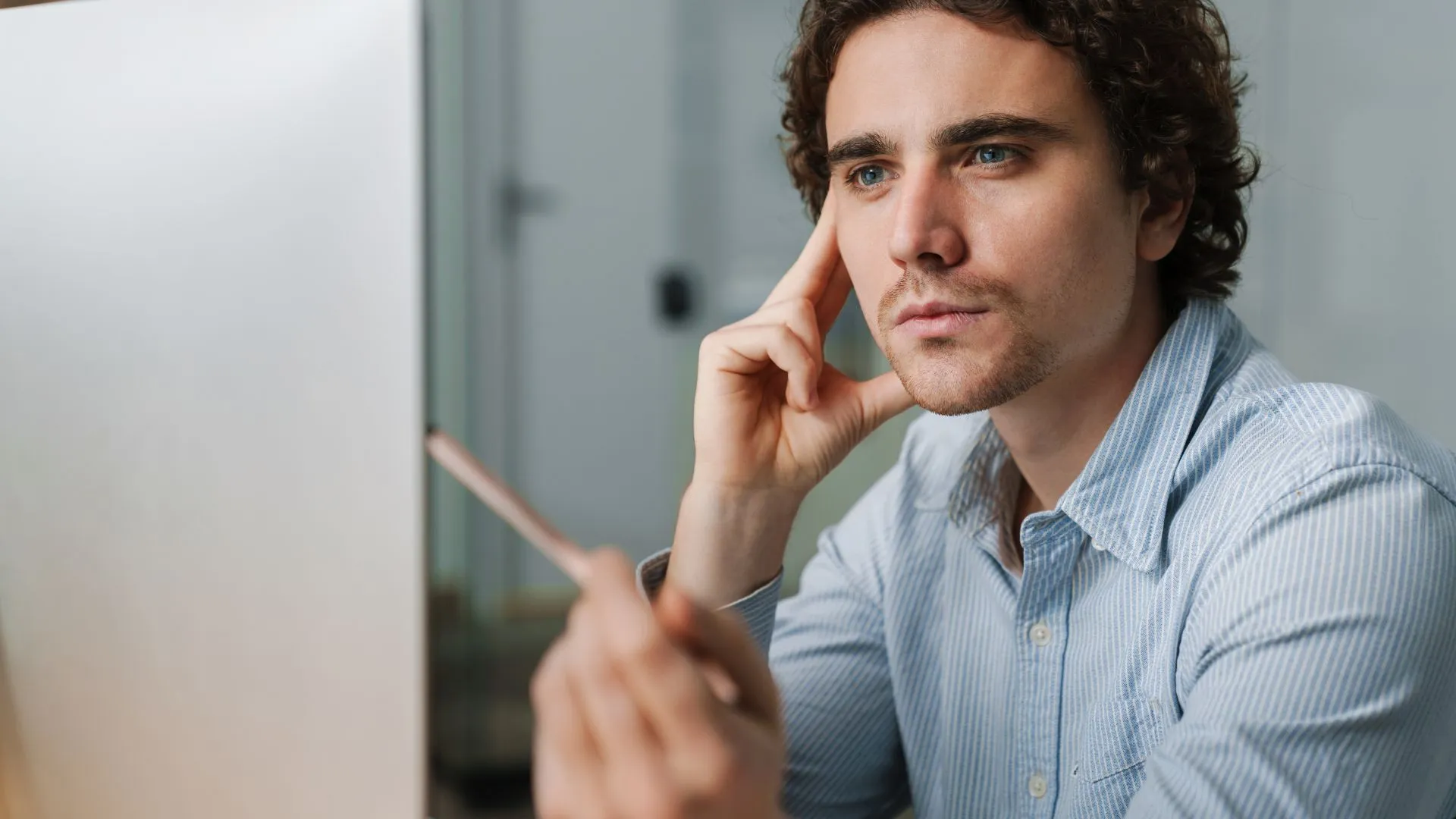 Young adult man in a light blue collared shirt sits at a desk, intently looking at his computer monitor with a pencil in hand—an expression of focused contemplation. The scene conveys the idea of ‘learning that works,’ with AI as a thinking partner to spark and support his problem-solving process.