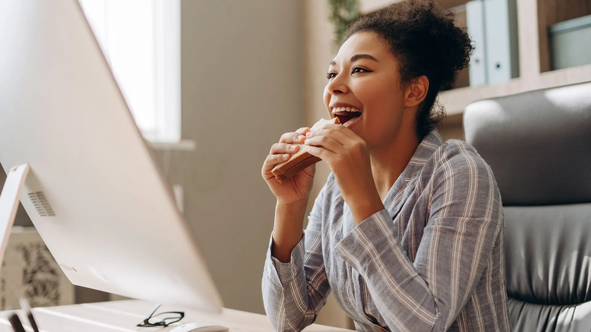 A smiling woman in a striped top takes a bite of a sandwich while seated at her desk, happily looking at her computer screen. This relaxed scene highlights how Electives is enjoyable for employees, fostering a positive and satisfying work experience.