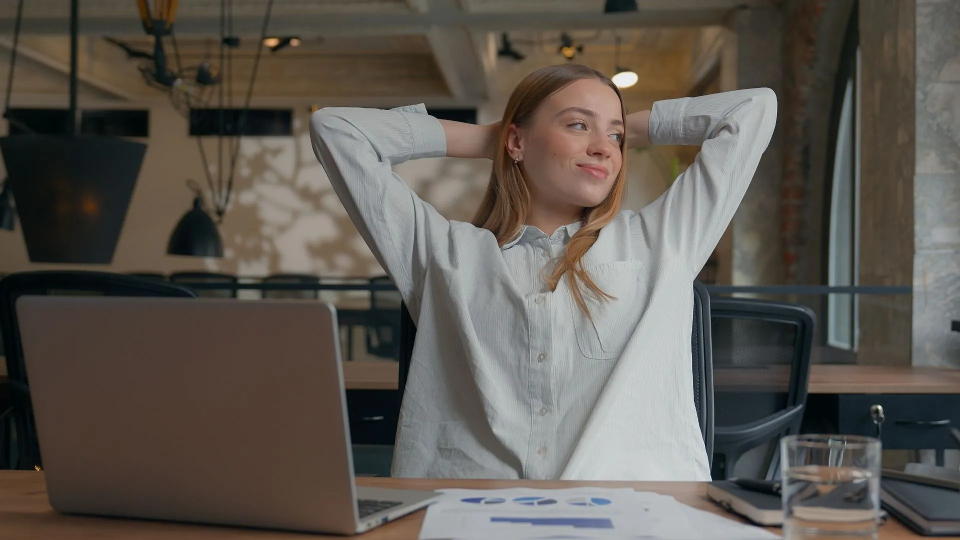 A woman in a button-down shirt leans back with her hands behind her head, looking relaxed and confident at her desk with a laptop and charts in front of her — representing how Electives is easy for HR to manage.