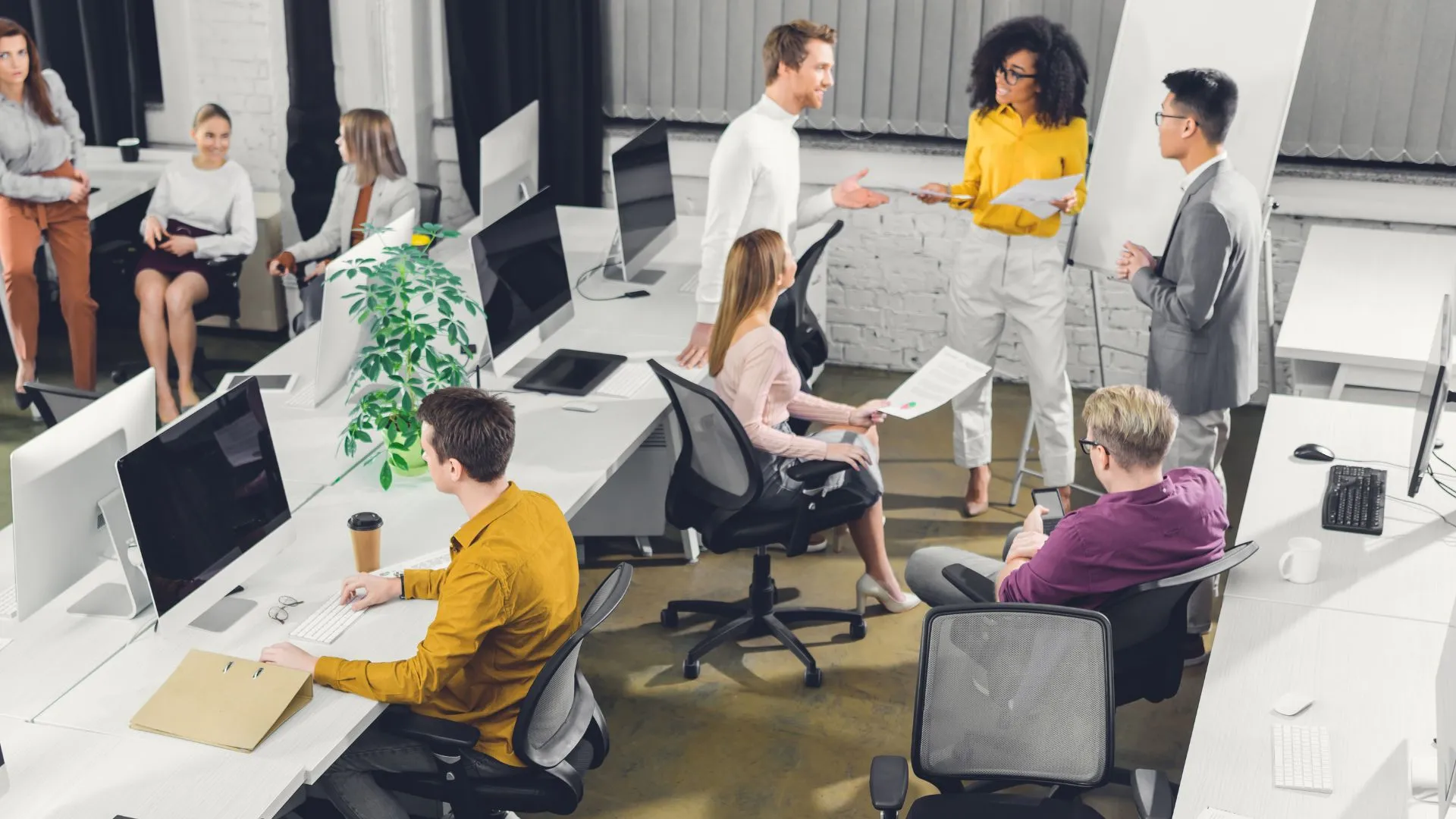 A busy open-plan office with multiple colleagues collaborating — some seated at desks, others standing and discussing documents. This dynamic setting highlights how Electives is effective for businesses, fostering seamless workplace learning and teamwork.