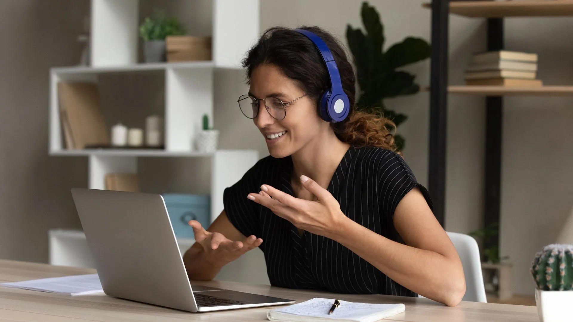 A woman is working from her home office wearing over ear headphones. She is talking to someone on her laptop, smiling and gesturing while she does so.