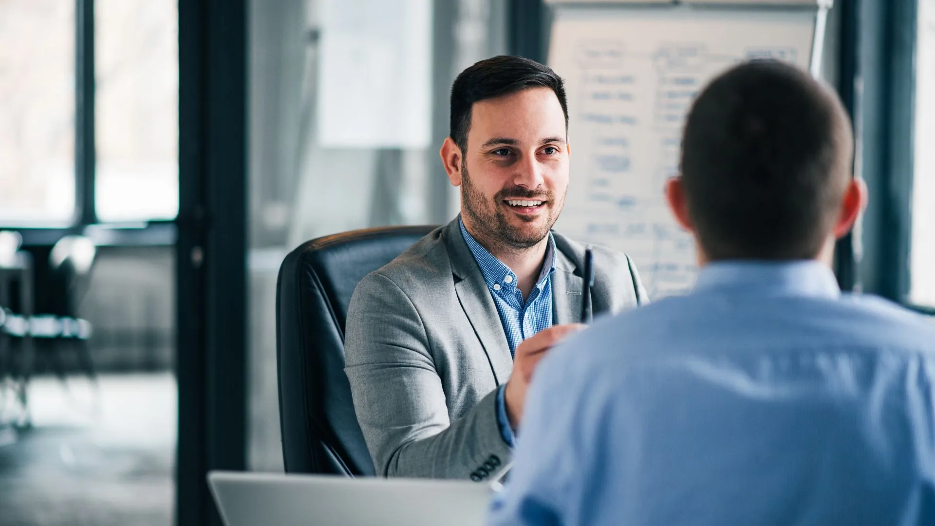 A manager is conducting a one on one meeting in the office with an employee. We are looking over the emplyee's shoulder at the manager's smiling face.