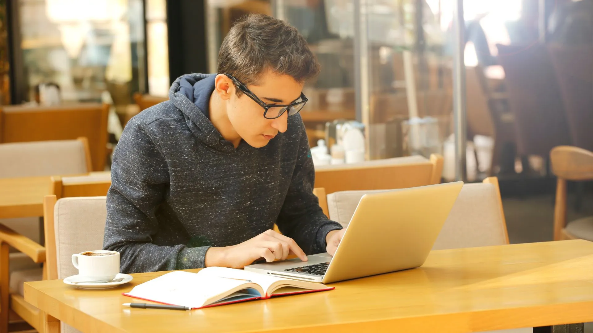 A man is sitting at a table in a diner on his laptop. There is an open notebook and a cup of coffee next to him.