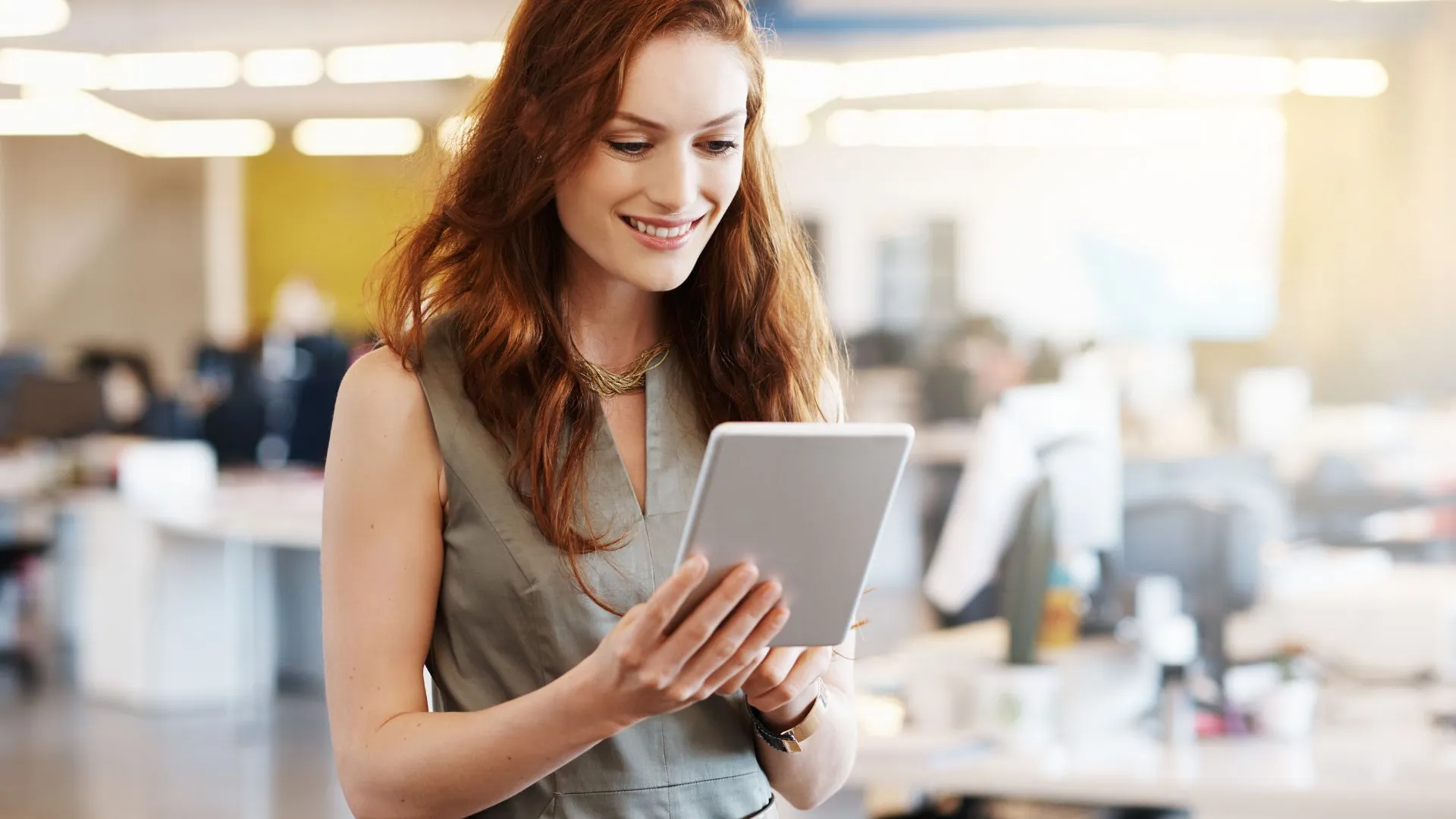 A woman is standing in an open office space, smiling at the tablet she's holding.