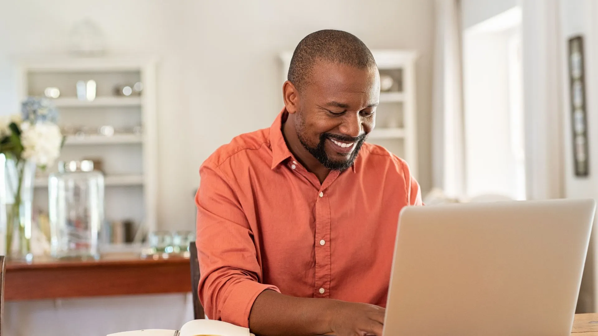 A man is sitting at his dining room table working on his laptop and smiling.