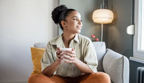 A young woman is sitting in a soft chair holding a cup of tea and smiling out the window