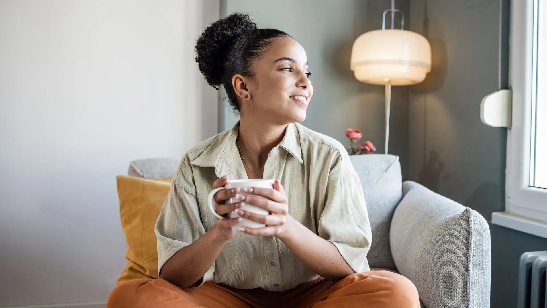 A young woman is sitting in a soft chair holding a cup of tea and smiling out the window