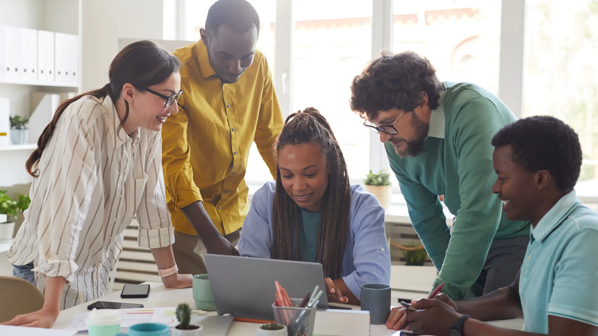 There are five people around a computer collaborating on a project. One woman is sitting down at the desk while everyone else is standing around her.