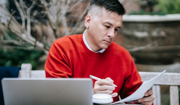 A man is working on his laptop outside. He is reviewing a document with a pen in hand.