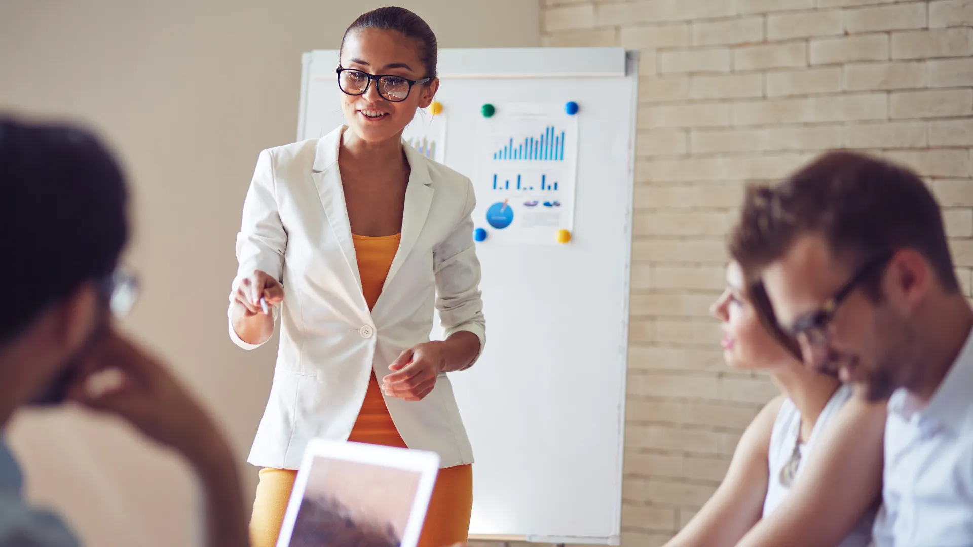 There's a woman standing in front of two people who are sitting down and she is presenting a chart that sits behind her.