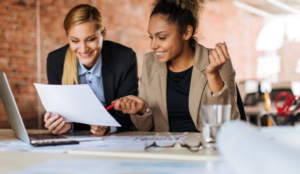 Two female-presenting coworkers are consulting with each other on a project and smiling.
