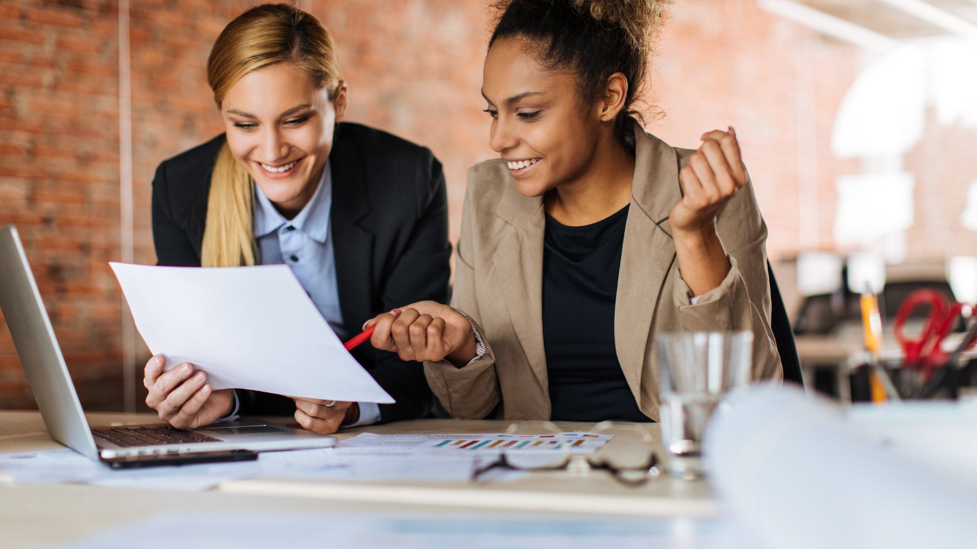 Two female-presenting coworkers are consulting with each other on a project and smiling.