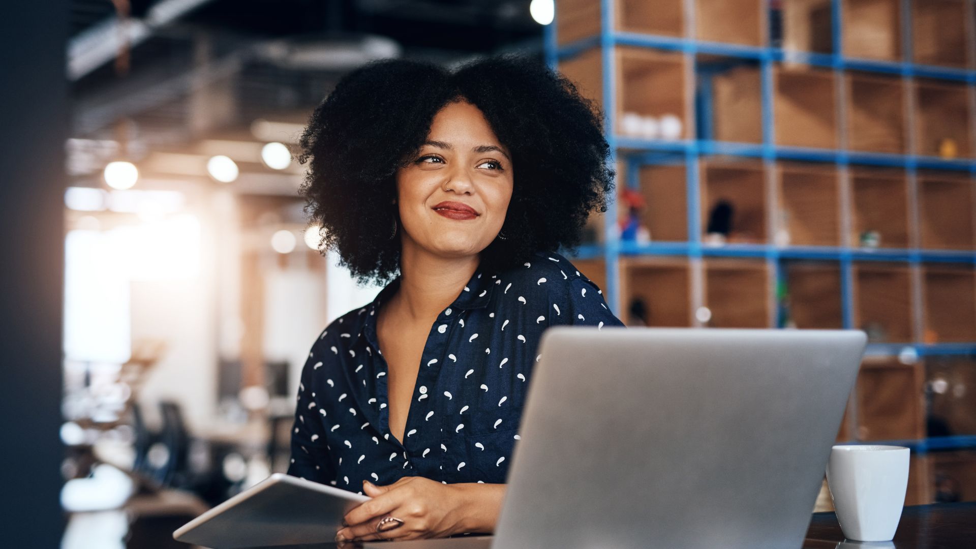 A woman is at her laptop in a common office area, looking and smiling off into the distance.