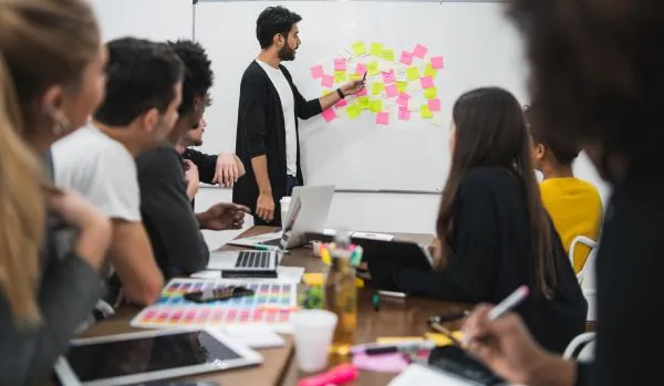 A diverse team of people are sitting around a conference table looking at their manager who is at the whiteboard discussing post-it notes that are stuck to it.