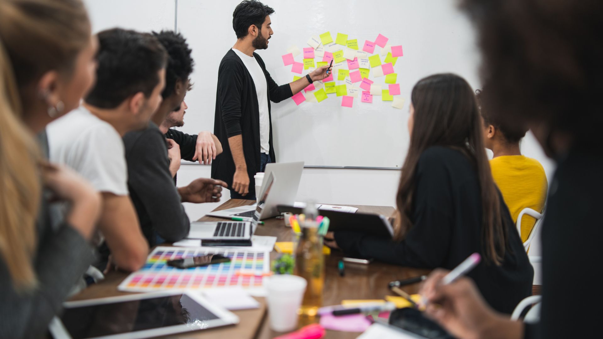 A diverse team of people are sitting around a conference table looking at their manager who is at the whiteboard discussing post-it notes that are stuck to it.