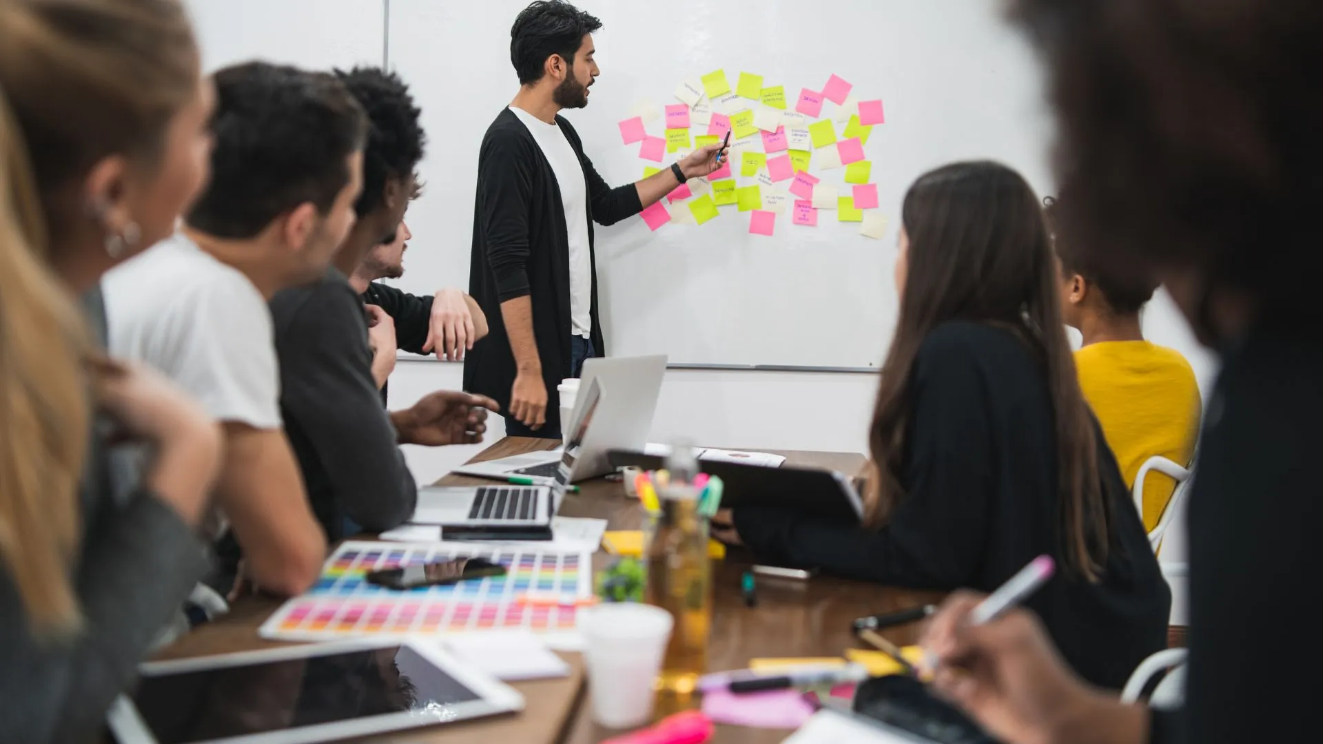 A diverse team of people are sitting around a conference table looking at their manager who is at the whiteboard discussing post-it notes that are stuck to it.