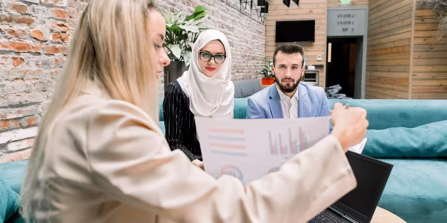 Three colleagues — a woman in a light blazer, a woman wearing a hijab, and a man in a blue jacket — sit on a sofa in a modern office, discussing a document with charts. The woman in front holds a paper with diagrams, pointing at the data, while the others listen attentively. In the background, a brick wall, plants, and contemporary interior details are visible.