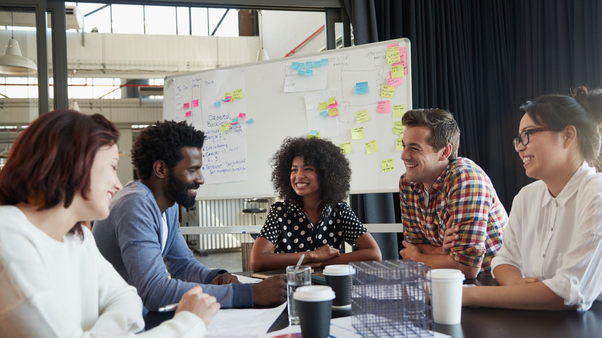 A diverse group of coworkers are smiling around a conference table with a whiteboard in the background.