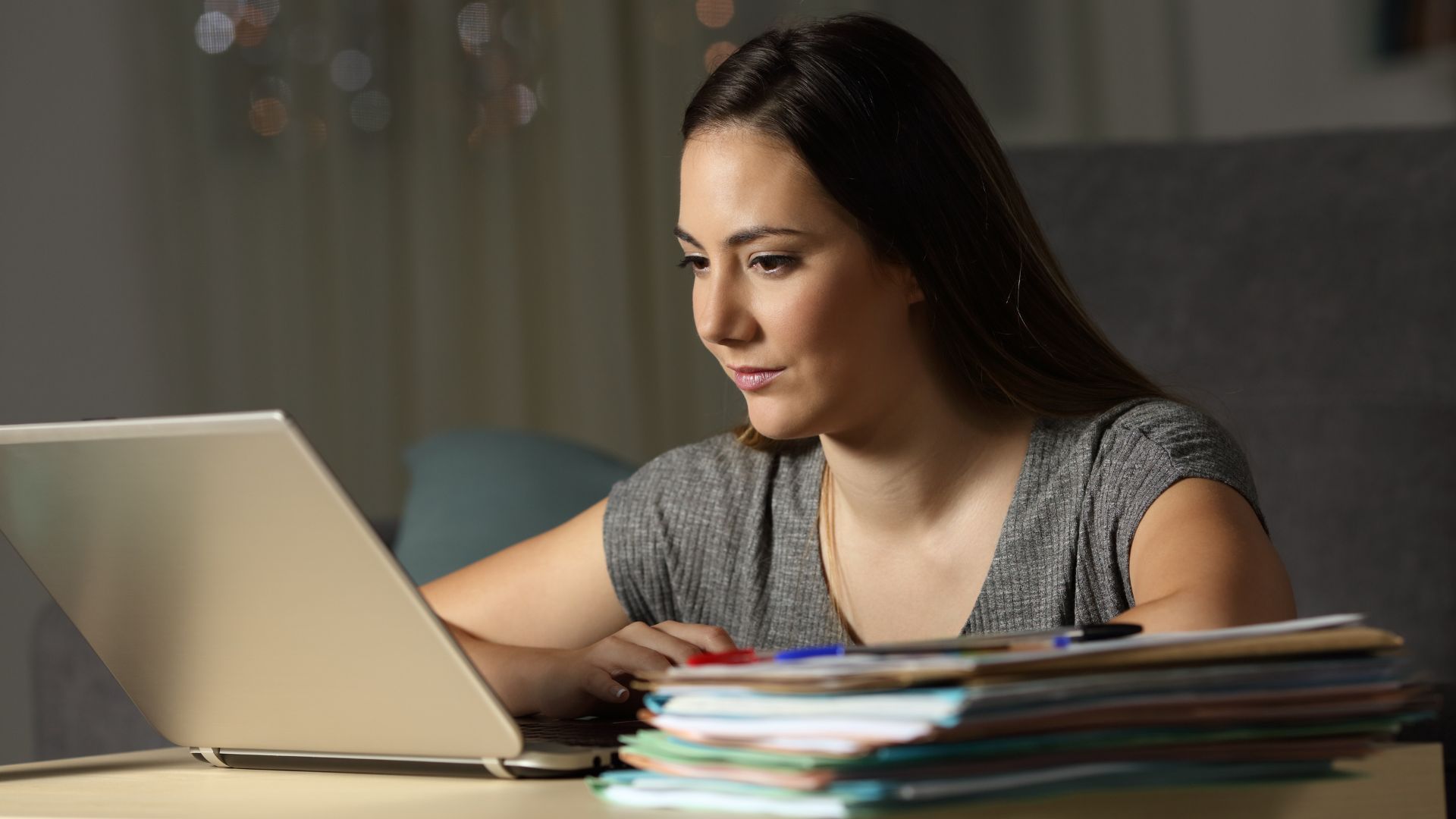 A woman is working on her laptop with a stack of paperwork next to her.