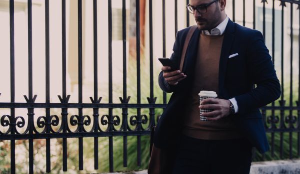 A man in a suit is looking at his phone while walking to work with coffee in his other hand.