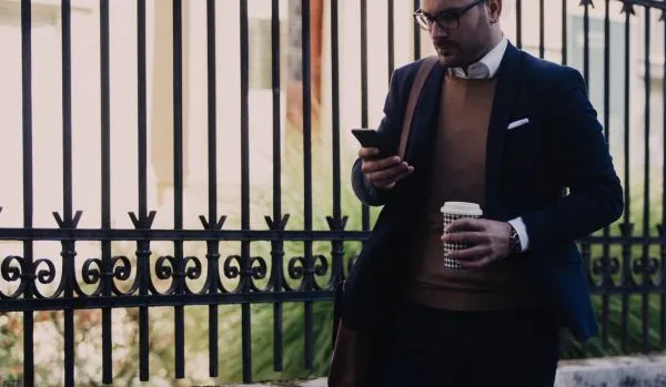 A man in a suit is looking at his phone while walking to work with coffee in his other hand.