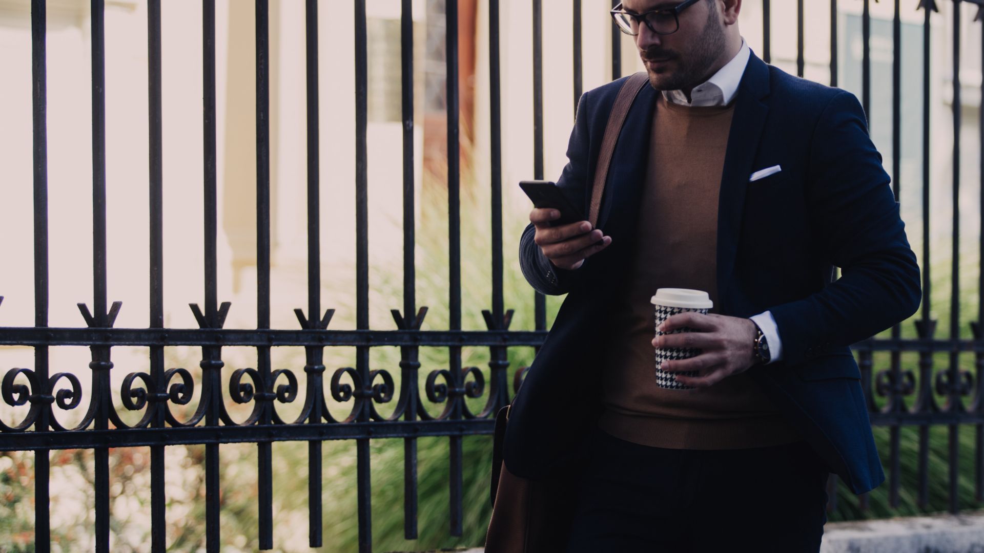 A man in a suit is looking at his phone while walking to work with coffee in his other hand.