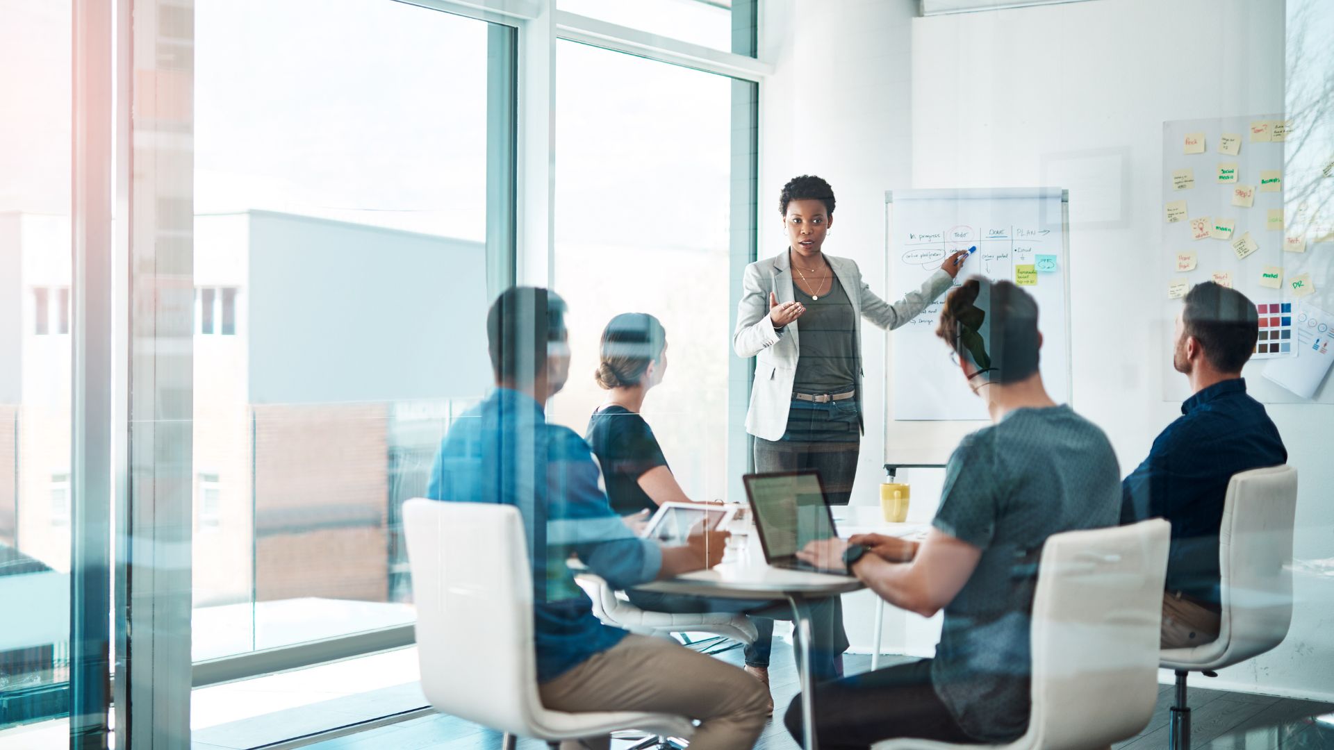 A female-presenting woman is standing at a flipchart at the head of a conference table leading a training for a diverse group of colleagues.