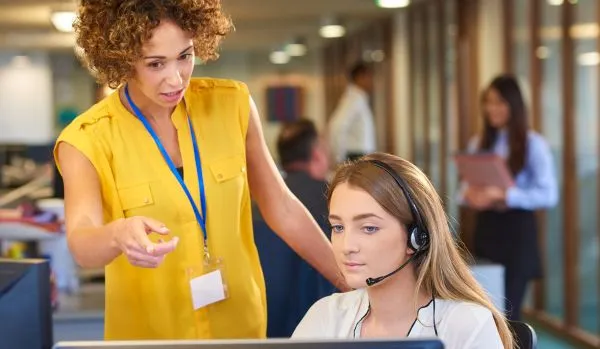 A manager is standing behind a seated employee, pointing at the computer screen and providing some on-the-job learning.