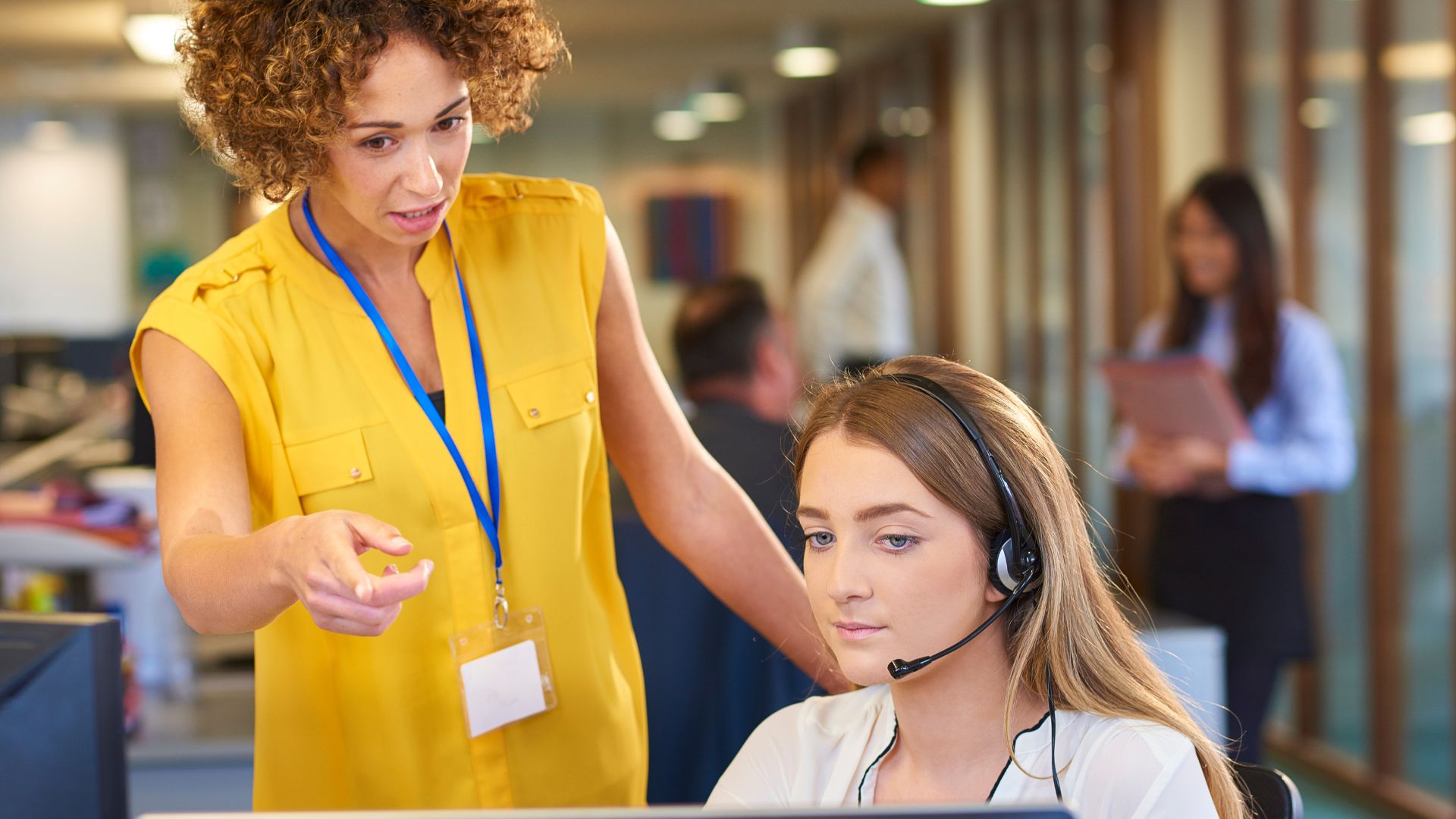 A manager is standing behind a seated employee, pointing at the computer screen and providing some on-the-job learning.