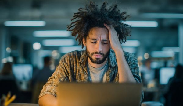 A manager is holding his head looking down at the laptop on his desk in the office.