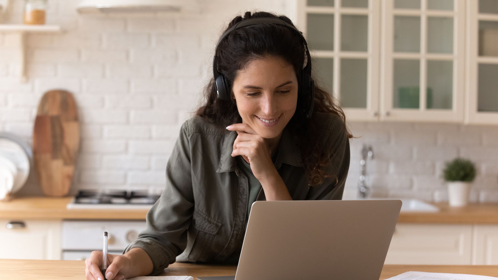 A woman is wearing headphones and smiling down at her laptop. She's working from home and taking notes from the webinar she's attending.
