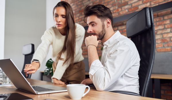A female-presenting manager is standing next to her male-presenting employee, teaching him how to do something on his laptop.