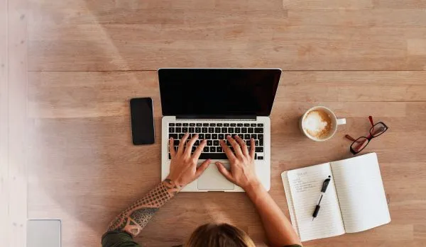 We're looking down from overhead of a person using their laptop. There is a notebook and coffee on the table next to them. 