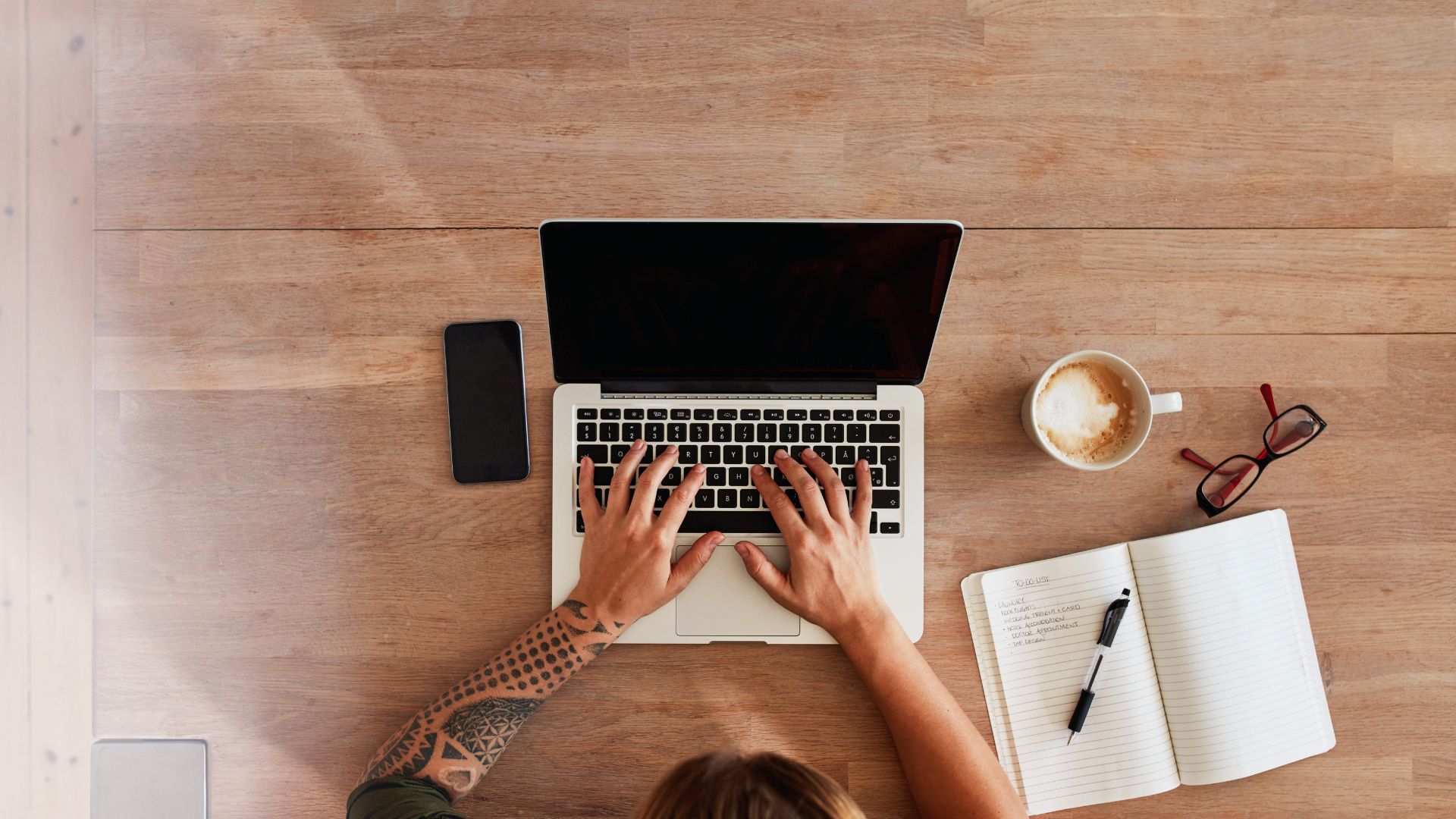 We're looking down from overhead of a person using their laptop. There is a notebook and coffee on the table next to them. 