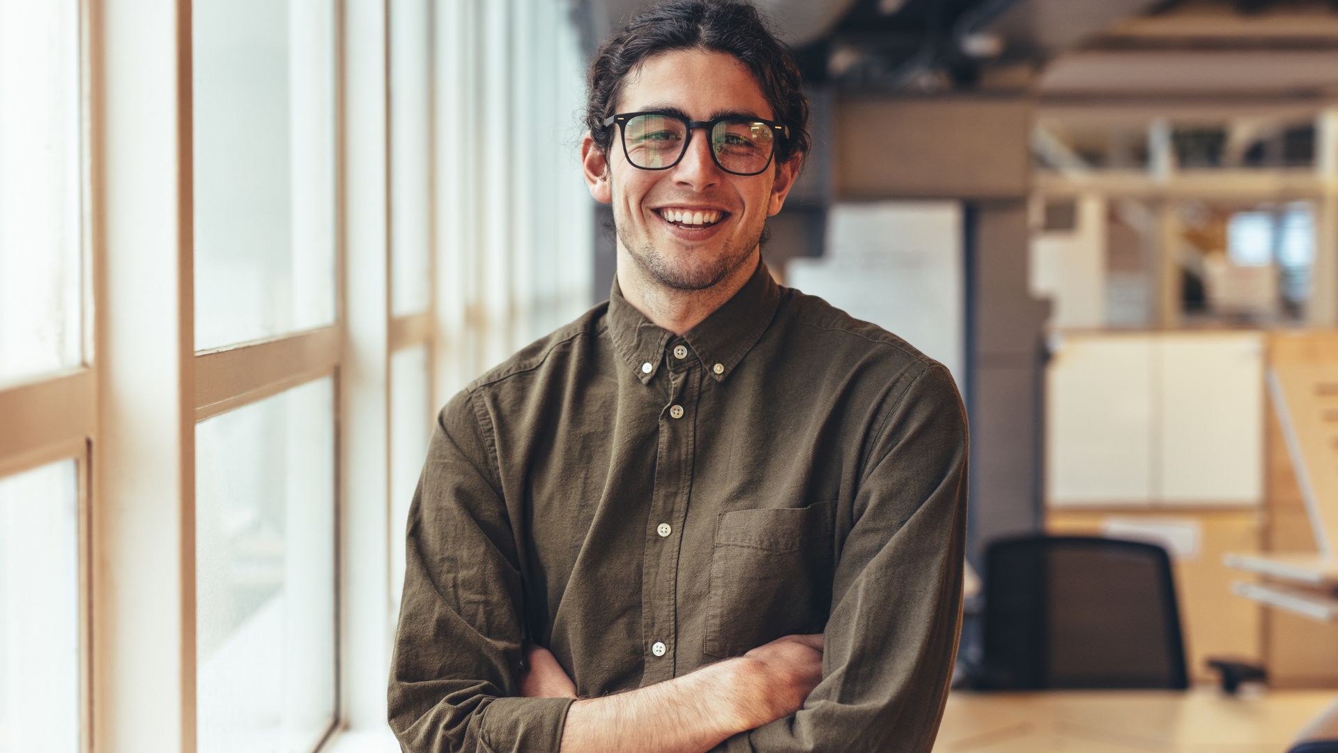 A smiling nonbinary person in glasses is standing in an open office space next to some windows.
